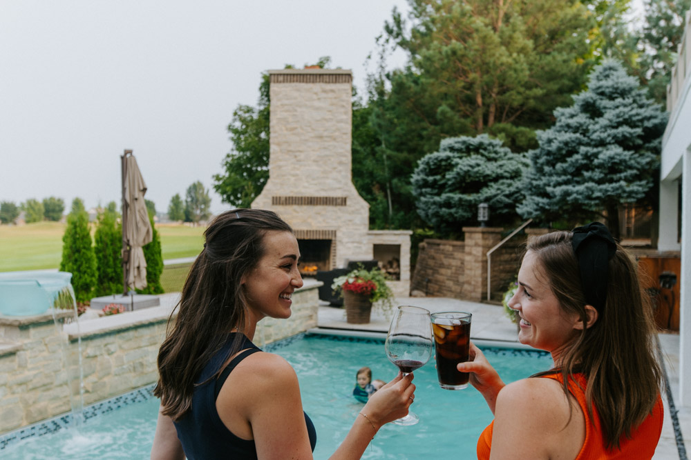 Two women clink glasses while relaxing by a backyard pool, with an outdoor fireplace and lush greenery in the background.