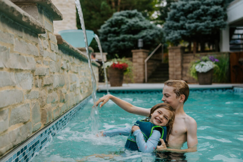 A young boy embraces his smiling younger sister as they play under a cascading water feature in a backyard pool.