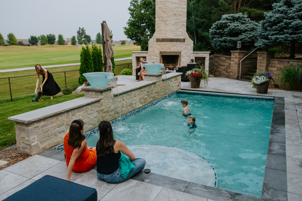 A luxurious backyard pool area features a small in-ground pool surrounded by stone and tile. Two women sit at the pool’s edge with their feet in the water, while two children swim nearby. A cascading water feature adds movement to the pool. In the background, a woman plays with a dog on a lush green lawn, and another woman sits near a large outdoor fireplace, where a man is also seated. The landscaping includes potted flowers, manicured shrubs, and a stone retaining wall, creating a cozy and elegant outdoor gathering space.